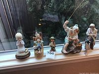 Photo showing six porcelain and ceramic figurines on a windowsill, including a praying child, two boys with animals, a fisherman, an older man with a pipe, and a woman knitting.