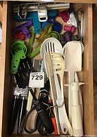 Top-down view of drawer with assorted kitchen gadgets including measuring spoons, spatulas, scissors, can openers, bag clips, and bottle stoppers.