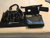 Top view of Makowsky handbag with fur accents, The Sak black textured shoulder purse, Jacqueline Ferrar black leather purse, and an additional small black pouch on beige surface.