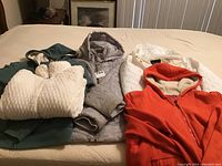 All four jackets displayed on bed showing variety of colors, styles, and fabrics: green fleece, gray quilted, white quilted, and orange fleece-lined hoodie.