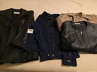 Multiple coats laid out together including two black leather coats, one blue denim jacket, and one taupe trench-style coat.