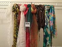 Photo of 7 scarves hanging on a rod, various colors and patterns including white lace, brown abstract, pink-red, green-orange floral, leopard print, turquoise floral, and large blue floral print scarf.
