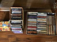 Overview photo showing the cardboard box filled with a large number of CDs and two woven baskets containing cassette tapes and additional CDs. A multi-disc CD collection titled 'Pop Memories of the '60s' is displayed.