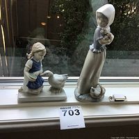 Two porcelain figurines placed on a windowsill, one girl feeding a goose and one standing girl with a basket