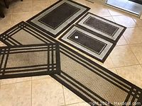 Four outdoor carpets laid on floor, one long runner with beige and black pattern, three smaller rugs with varying patterns and colors in dark brown and beige. Flooring is tile.