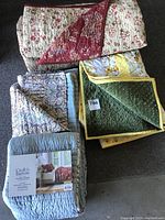 Photo showing all five quilts folded with visible colors and patterns including blue Croft & Barrow quilt