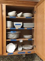 Full view of four shelves inside wooden cabinet holding various white ceramic dishes organized and labeled by type and quantity.