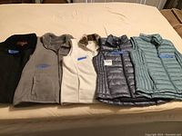 Five women's vests laid out on bed, showing color and style variety including fleece and quilted fabric with different collar styles.