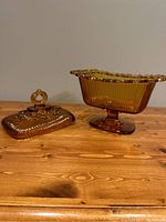 Side view of amber Indiana Glass candy dish with lid removed, showing the footed base and ribbed vertical lines on the bowl, and the lid placed beside it with a loop handle and floral detail.
