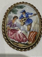 Front close-up view of the antique Limoges brooch showing the courting couple hand-painted scene and gold-tone braided frame.