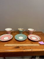 Front view of 3 Aynsley tea cups and matching saucers, two pink and gold, one mint green and gold, arranged side by side on wooden table with ruler for scale.