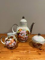 Three-piece vintage Bavaria porcelain tea set displayed together on wooden surface: coffee pot, cream jug, and lidded sugar bowl with iridescent finish and floral pattern with silver accents.