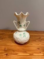 Full front photo of off-white Belleek vase with doubled harp-shaped handles, scalloped top, raised relief pattern, and green hand painted shamrocks on body and neck.