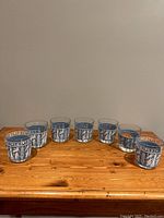 Set of seven blue glass drinking glasses each decorated with white Grecian figures and patterns, arranged side-by-side on wooden surface.