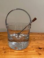 Front view of ice bucket with ice tongs resting inside, showing clear glass construction, thick base with suspended bubble, chrome handle, and original label.