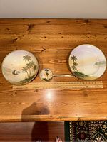 Top-down view of the set on wooden table with ruler indicating size; shows hand-painted bowl, plate, and spoon with tropical palm tree design and gold trim.