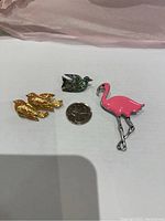 Clear image of the three vintage animal brooches arranged on a white background with a quarter for scale: a silver tone and pink enamel flamingo, a gold tone and peach enamel pair of birds, and a bird with abalone shell inlay.
