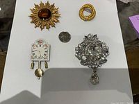 Four vintage brooches arranged with a quarter for size comparison on white paper. Includes sunburst brown glass gold tone brooch, gold tone loop brooch, crystal silver tone brooch with drop, and clock-shaped brooch.