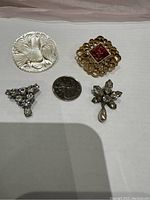 Four vintage brooches placed on a white background alongside a quarter for size comparison. Includes a carved Mother of Pearl bird brooch, gold-tone agate stone brooch, silver-tone rhinestone and pearl brooch, and silver-tone rhinestone cluster brooch.