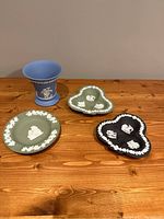 Photo showing four Wedgwood ceramic pieces on wooden table: one blue vase, one black club-shaped dish, one green club-shaped dish, and one round green dish, all with white relief decoration.