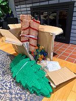 Unassembled green tinsel palm leaf sections, red and white tinsel trunk pieces inside a partially open cardboard box with multi-colored G40 LED lights visible and assembly instructions.