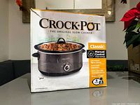 Box front showing Crock-Pot Classic Slow Cooker with food inside and text about manual control and features. Box is damaged with visible tears and bends.