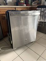 Full view of the dishwasher placed on a tiled floor. Stainless steel exterior with visible rubbed areas on the door.