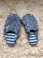 Pair of grey fuzzy slippers with striped insoles and small bows, placed on a beige shaggy rug.