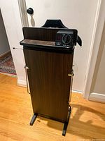 Full view of the pant press standing on wooden floor against white walls, showing the full height vertical structure with wood-grain finish, padded press section, and metal support bars.
