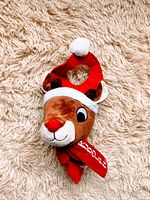 Photo of a Rudolph-themed doorknob cover placed on a textured cream rug, showing details of the plush red nose, brown face, Santa hat, and scarf.