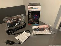 Photo shows the boxed Projection Plus Christmas projection light, the black projector unit wrapped in plastic, power cord with attached warning tag, and instruction manual.