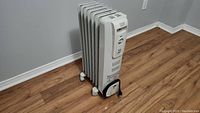 Side view of white oil-filled radiator heater standing on wood floor, showing fins and wheels.
