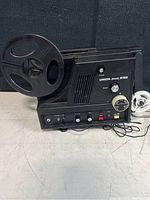Front and side view of black Chinon Sound 6100 Super 8 film projector showing the large reel, various control knobs, and power cord.