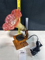 Miniature Victrola styled lamp shown from front with measuring tape indicating size approximately 12 inches tall. Wooden base with black turntable and crank handle, multicolored pink glass flower lamp shade.
