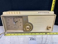 Front view of a vintage beige Motorola clock radio featuring an analog clock face, five white buttons on top, a tuning dial, and a small knob. A ruler indicates dimensions around the radio.