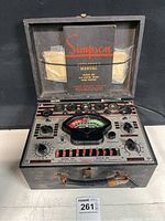 Simpson Model 305 tube tester opened in metal case showing control panel and meter with labeled dials and tube sockets; manual tucked in case lid.