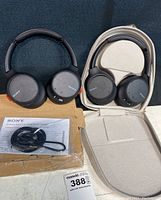 Two black Sony wireless noise cancelling headsets side by side. One headset outside case with cables and manual in front.