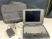 Photo showing vintage Compaq Contura laptop open with visible keyboard, screen, and trackball, with black carrying bag behind and wired mouse and adapter cables in front.