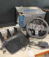 View of Logitech G920 wheel, pedal set, power adapter, and original box with visible wear