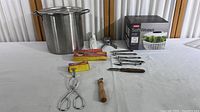 Wide shot showing large stainless steel Vollrath pot, peelers, tongs, wooden handled kitchen tool, and boxed OXO SoftWorks steamer basket.