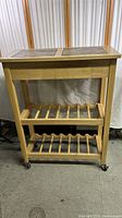 Full view of the wooden rolling wine rack showing overall structure, slatted shelves, casters, and tile top.