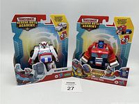 Two Transformers Rescue Bots Academy action figures in original packaging. Left: Autobot Ratchet in white, red, and blue. Right: Optimus Prime in red, blue, and white. Both labeled 2-in-1 transformation.