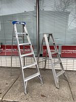 Two metal step ladders standing outside near a window, one taller with blue plastic caps and the other shorter, both showing signs of wear.