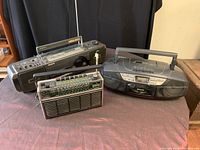 Three vintage electronic devices on table: Sony boombox, Panasonic portable CD player, and Telefunken shortwave radio.