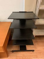 Front view showing all four shelves of the black metal audio stand and the rectangular base with caster wheels.