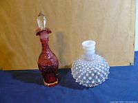 Photo of two decorative glass bottles, cranberry glass decanter with stopper on left and clear hobnail glass bottle on right, with beige background.