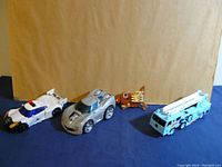 Four vintage Transformers toys arranged on blue cloth with brown cardboard background showing a variety of vehicle forms and a small robot figure.