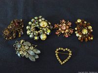 Six vintage brooches displayed on a dark surface; various styles including flowers and heart shapes, with rhinestone and faux pearl embellishments.