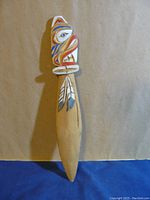 Front view of indigenous wooden letter opener showing the carved and painted totemic figure and feathers.