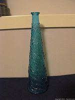 Large tall blue glass bottle with textured swirled pattern standing on a dark surface against a beige wall.